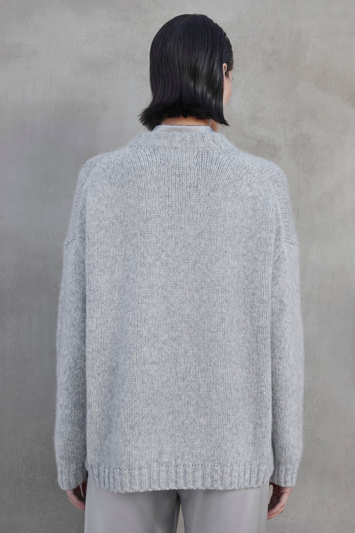   A person with short dark hair facing away, wearing an oversized CAMBRIA ALPACA SWEATER by Velvet by Jenny Graham and light-colored pants, standing against a plain gray wall. 