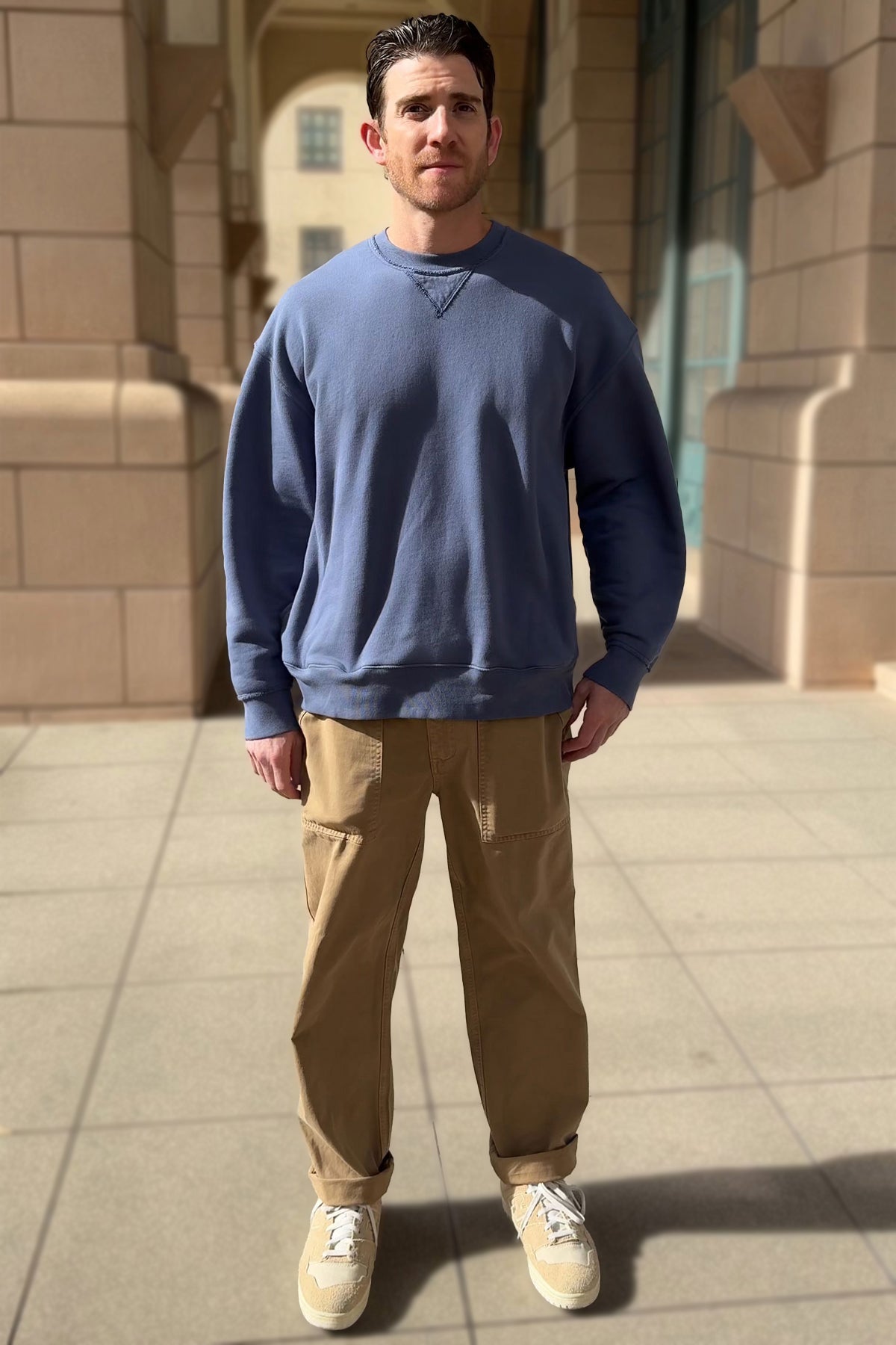   A man stands on a sunlit, tiled walkway wearing the FORREST SWEATSHIRT by Velvet by Graham & Spencer, paired with tan pants and beige sneakers, against a backdrop of stone walls and windows. 