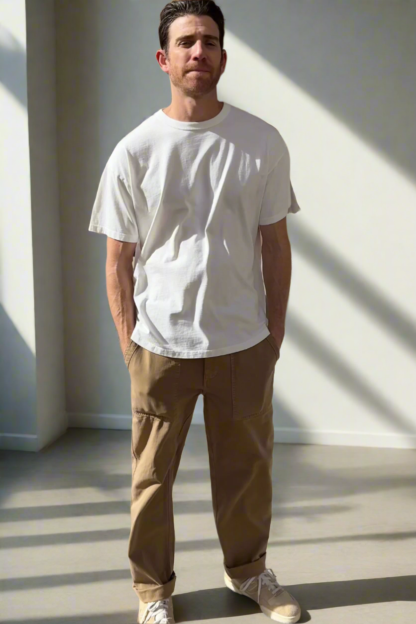 In a sunlit room, a man highlights everyday versatility by wearing the BRYAN TEE from Velvet by Graham & Spencer, crafted from premium pima cotton. He pairs it with brown pants and sneakers, displaying a relaxed fit that exudes casual ease as he comfortably tucks his hands in his pockets.-38325910634689