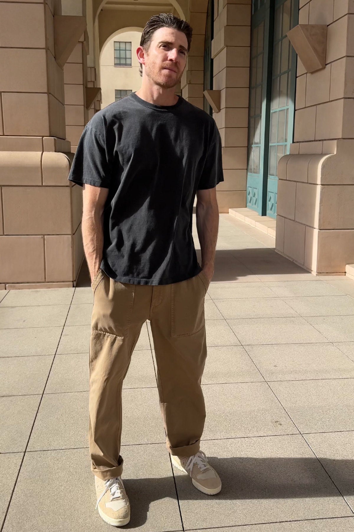   A man is outdoors in a casual ensemble, featuring the BRYAN TEE by Velvet by Graham & Spencer—a relaxed fit black T-shirt crafted from structured pima cotton—complemented by tan pants and beige sneakers, showcasing everyday versatility as he stands before a building with columns. 