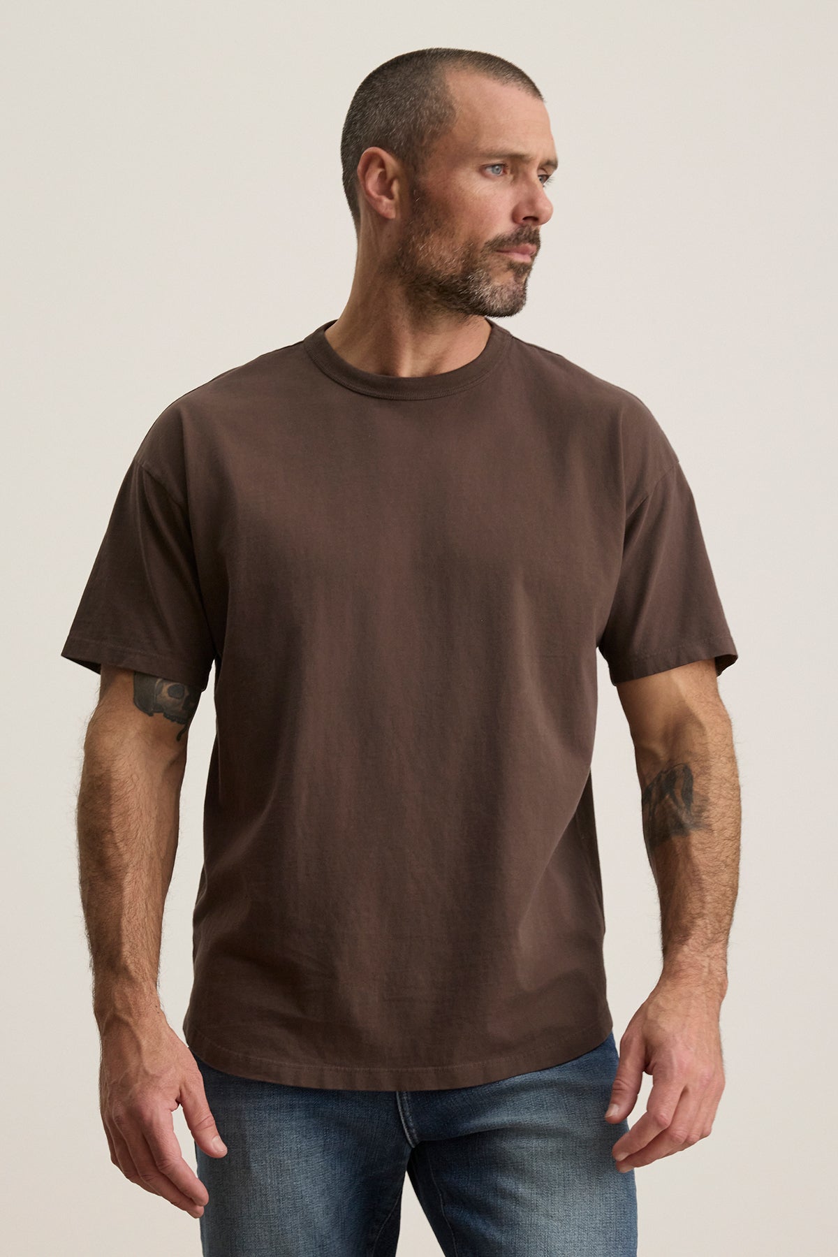   A tattooed man with a beard models the BRYAN TEE by Velvet by Graham & Spencer. This relaxed fit, structured pima cotton brown t-shirt is paired with jeans. He gazes left against a plain backdrop, embodying versatile and effortless everyday style. 