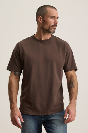 A tattooed man with a beard models the BRYAN TEE by Velvet by Graham & Spencer. This relaxed fit, structured pima cotton brown t-shirt is paired with jeans. He gazes left against a plain backdrop, embodying versatile and effortless everyday style.