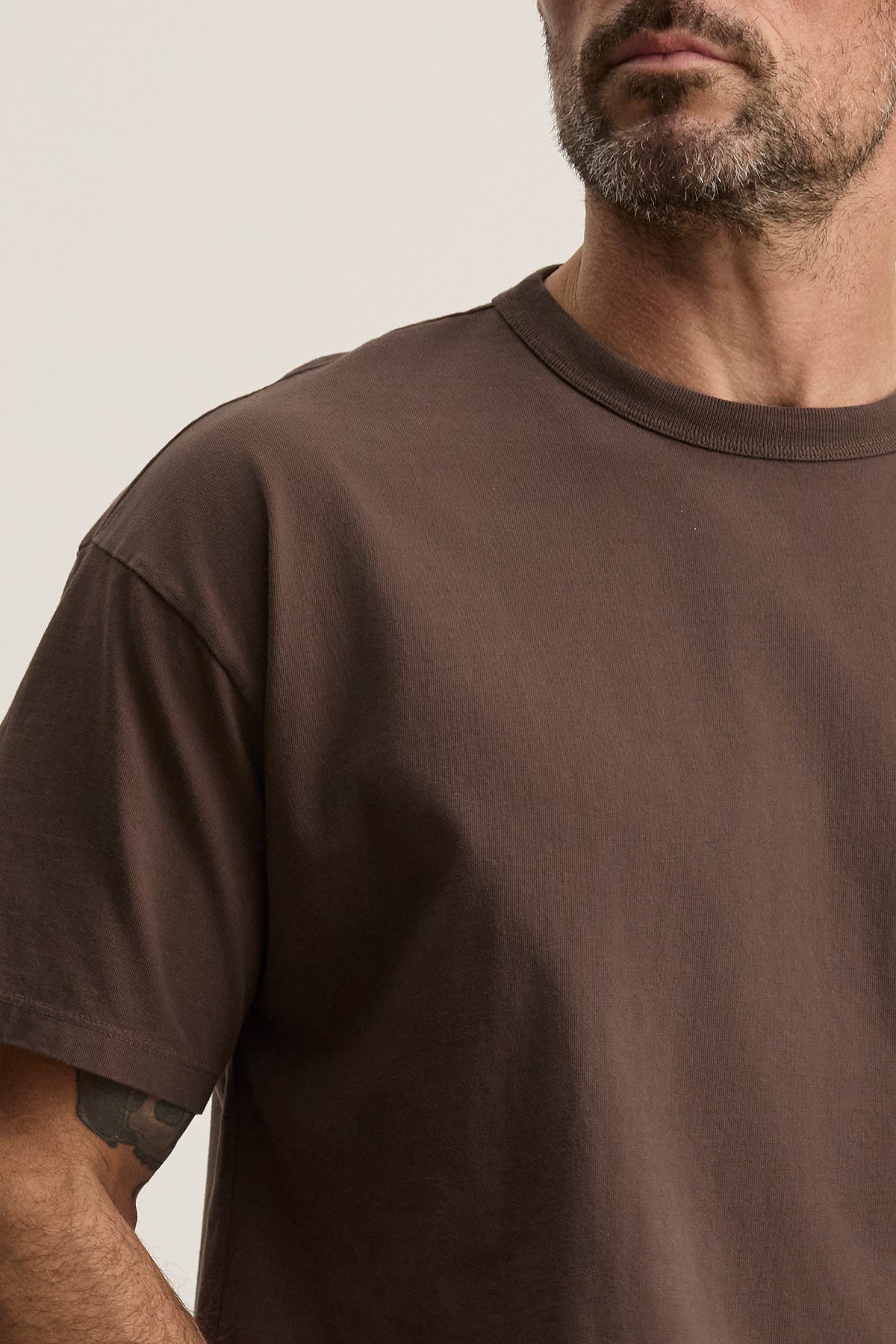 A man with facial hair, partially visible against a neutral background, exudes everyday versatility while wearing the Velvet by Graham & Spencer's relaxed-fit BRYAN TEE in brown.-38500086415553