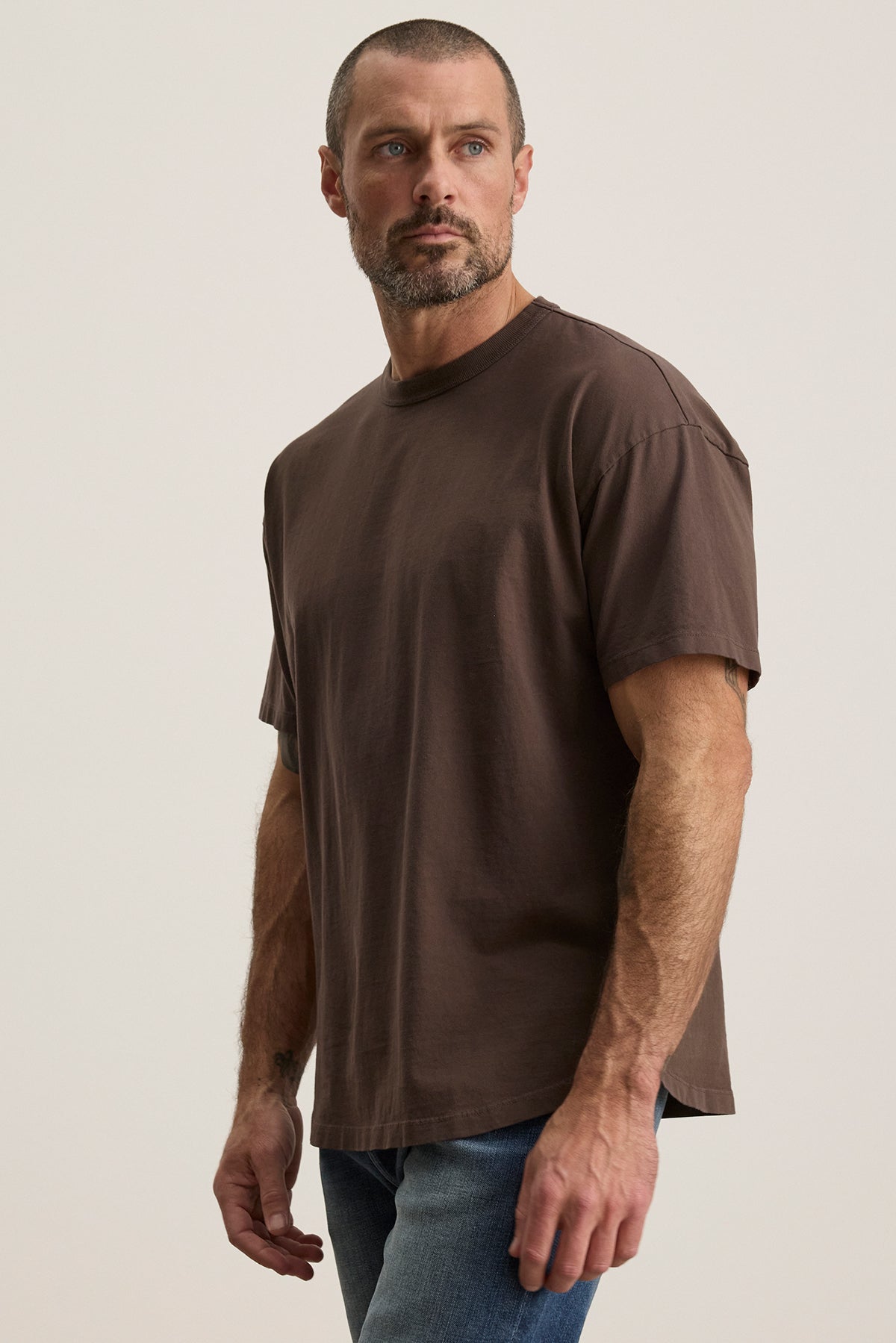   A bearded man wearing Velvet by Graham & Spencer's relaxed fit BRYAN TEE in brown structured pima cotton and jeans stands against a plain backdrop, highlighting everyday versatility. 