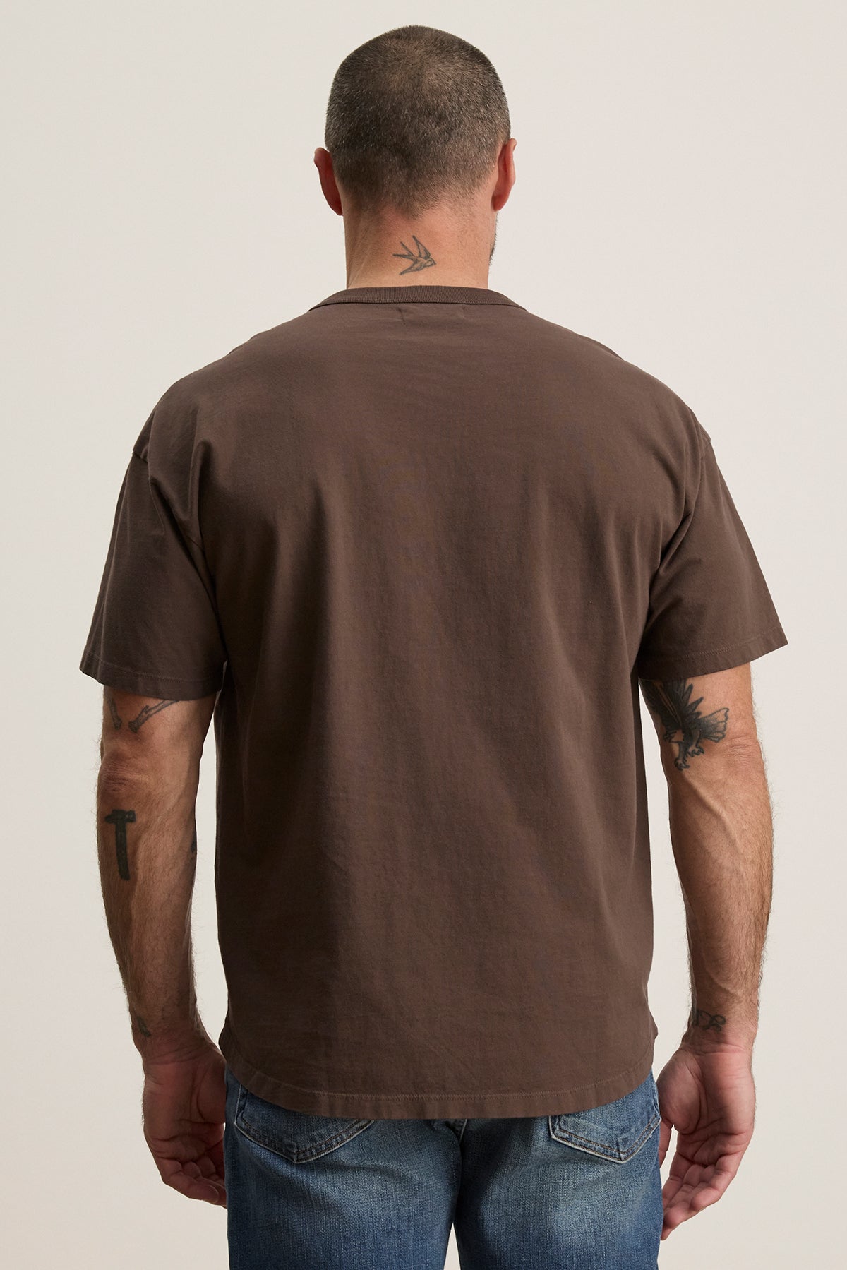   A man with short hair and tattoos displays everyday versatility wearing the brown BRYAN TEE by Velvet by Graham & Spencer, crafted from structured pima cotton, paired with relaxed-fit blue jeans, standing with his back to the camera. 