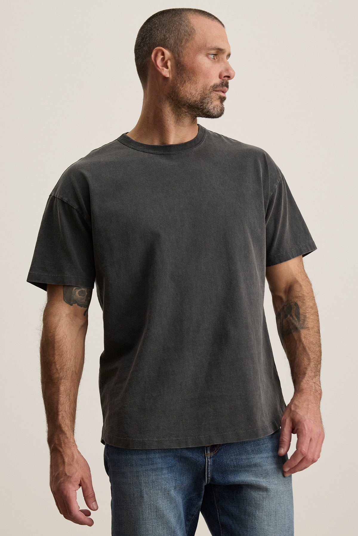 A man with short hair and a beard dons the plain dark BRYAN TEE from Velvet by Graham & Spencer, made of pima cotton for everyday versatility, paired with relaxed-fit jeans. He stands against a light background, gazing to the side.-38500076257473