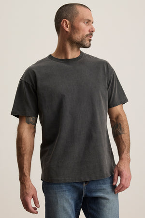 A man with short hair and a beard dons the plain dark BRYAN TEE from Velvet by Graham & Spencer, made of pima cotton for everyday versatility, paired with relaxed-fit jeans. He stands against a light background, gazing to the side.