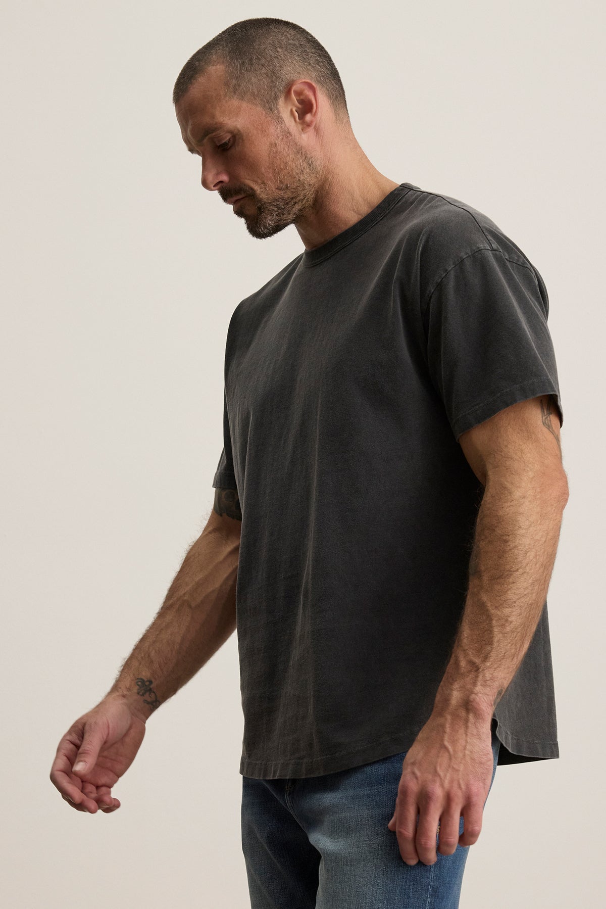   A man with short hair and a beard wears the relaxed fit BRYAN TEE from Velvet by Graham & Spencer, in faded black pima cotton, paired with jeans against a plain background. 