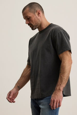 A man with short hair and a beard wears the relaxed fit BRYAN TEE from Velvet by Graham & Spencer, in faded black pima cotton, paired with jeans against a plain background.