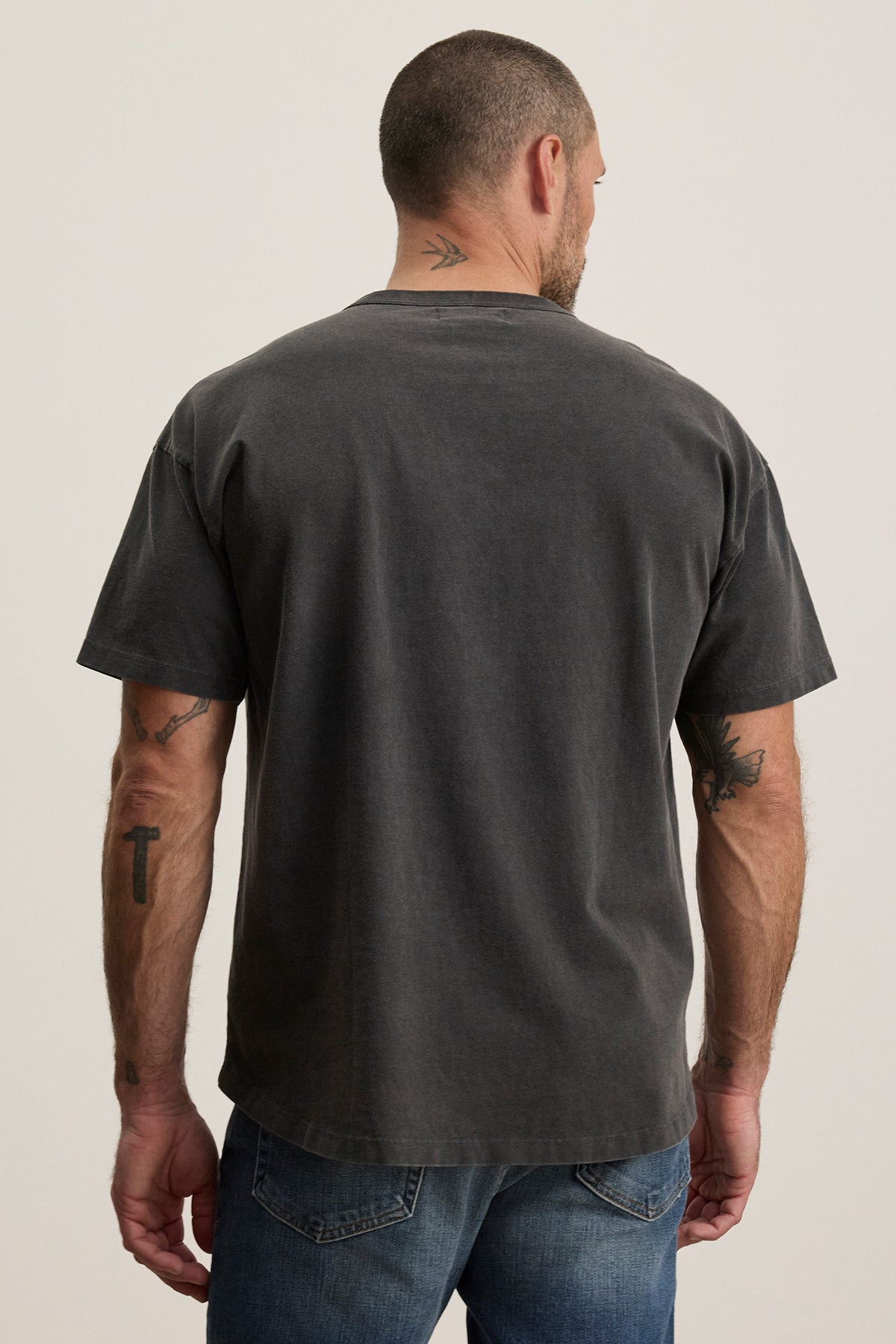   A man in a faded black BRYAN TEE by Velvet by Graham & Spencer, made from soft pima cotton, pairs with blue jeans. The relaxed fit highlights his arm and neck tattoos, showcasing everyday versatility. 