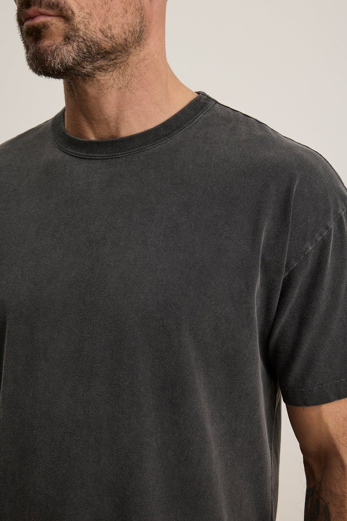   A man wearing the BRYAN TEE by Velvet by Graham & Spencer, made of luxurious pima cotton and featuring a faded black color, stands against a plain background. His face is partially out of the frame, highlighting the tee's relaxed fit and everyday versatility. 