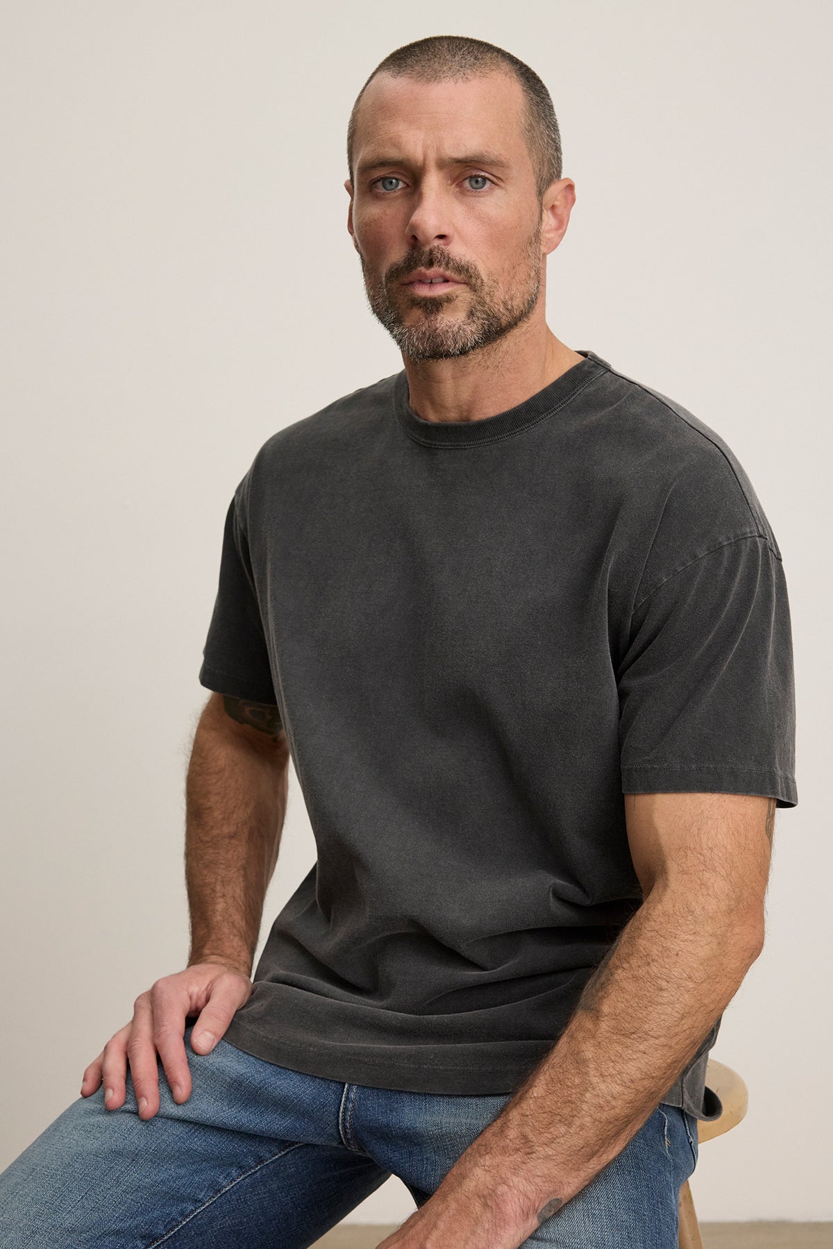   A man with short hair and a beard sits on a stool, wearing the BRYAN TEE by Velvet by Graham & Spencer, paired with blue jeans. The dark gray relaxed-fit T-shirt showcases everyday versatility against a plain background. 