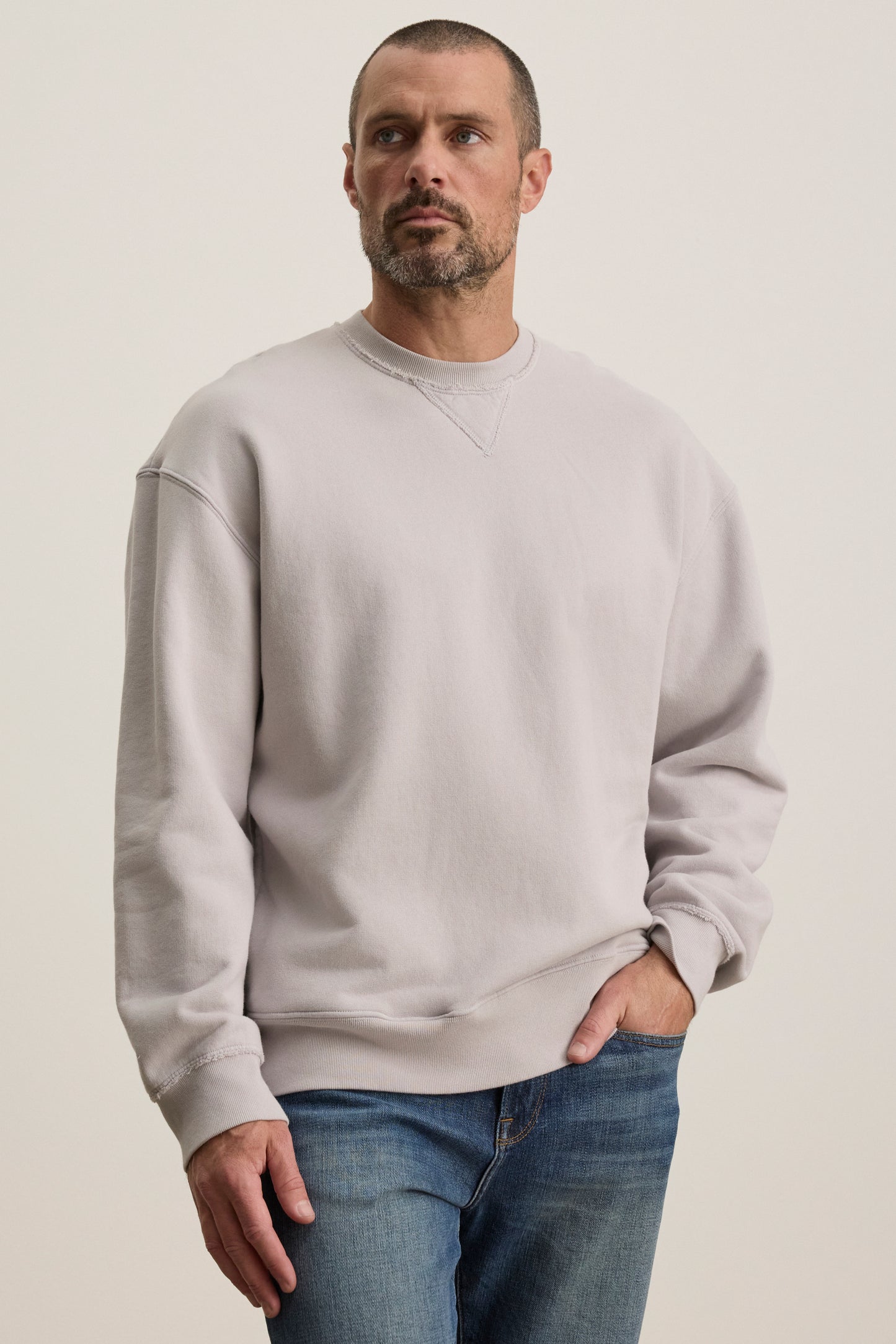 A man wearing a Velvet by Graham & Spencer FORREST SWEATSHIRT in relaxed-fit, light gray cotton fleece and blue jeans stands with one hand in his pocket against a plain background.-38500140843201