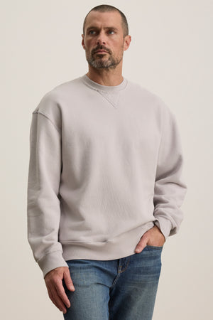 A man wearing a Velvet by Graham & Spencer FORREST SWEATSHIRT in relaxed-fit, light gray cotton fleece and blue jeans stands with one hand in his pocket against a plain background.