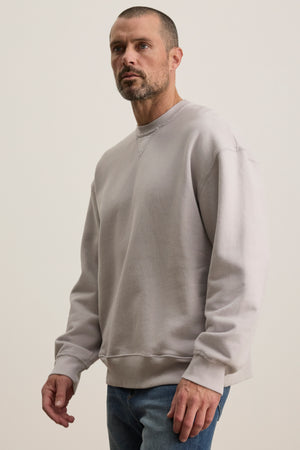 A person wearing the Velvet by Graham & Spencer FORREST SWEATSHIRT in a relaxed fit, made of gray cotton fleece, and blue jeans stands against a plain background, looking to the side.