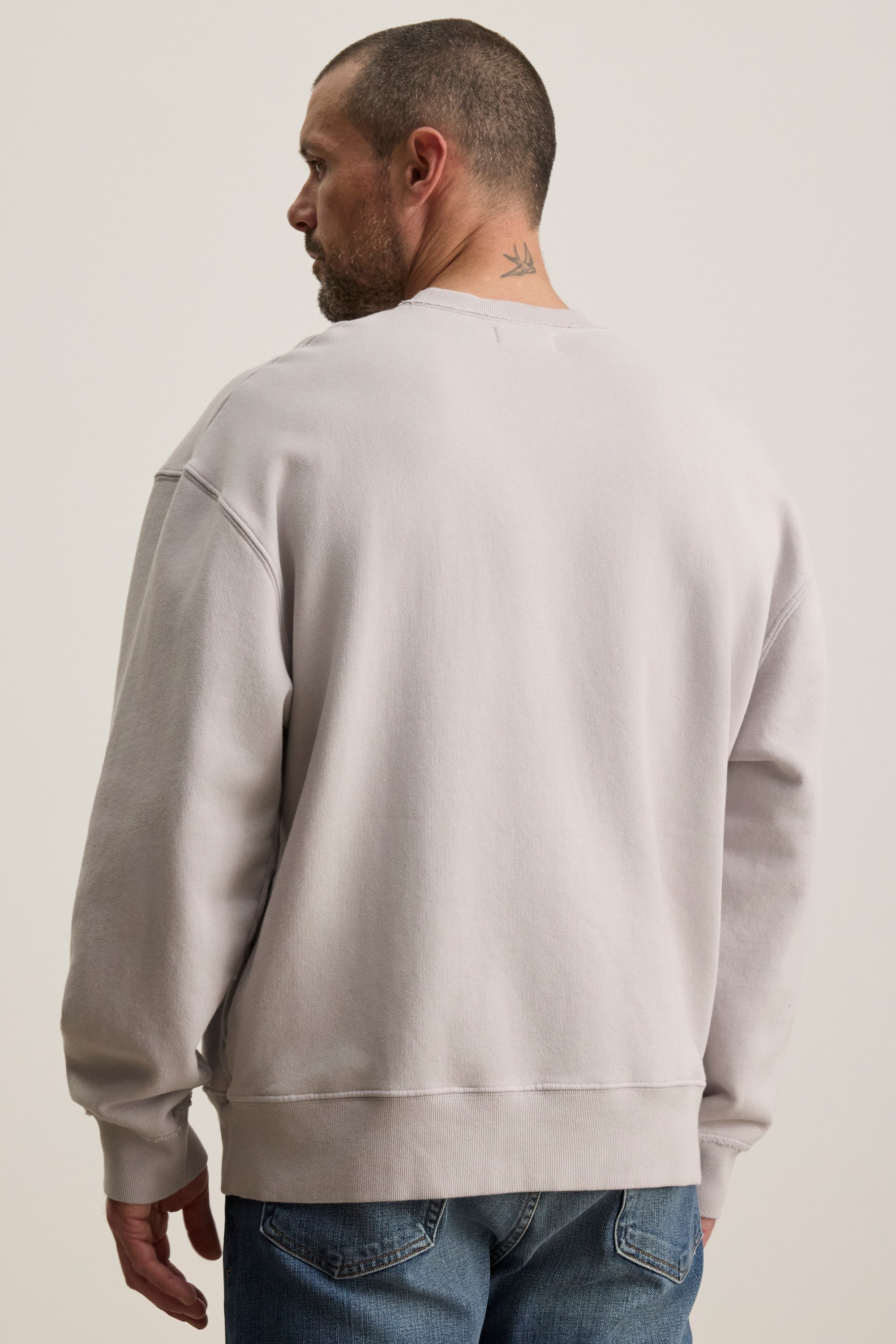   A man with short hair and a neck tattoo wears the light gray FORREST SWEATSHIRT by Velvet by Graham & Spencer, paired with relaxed-fit jeans, as he looks to the left. 