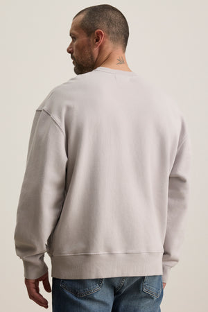 A man with short hair and a neck tattoo wears the light gray FORREST SWEATSHIRT by Velvet by Graham & Spencer, paired with relaxed-fit jeans, as he looks to the left.