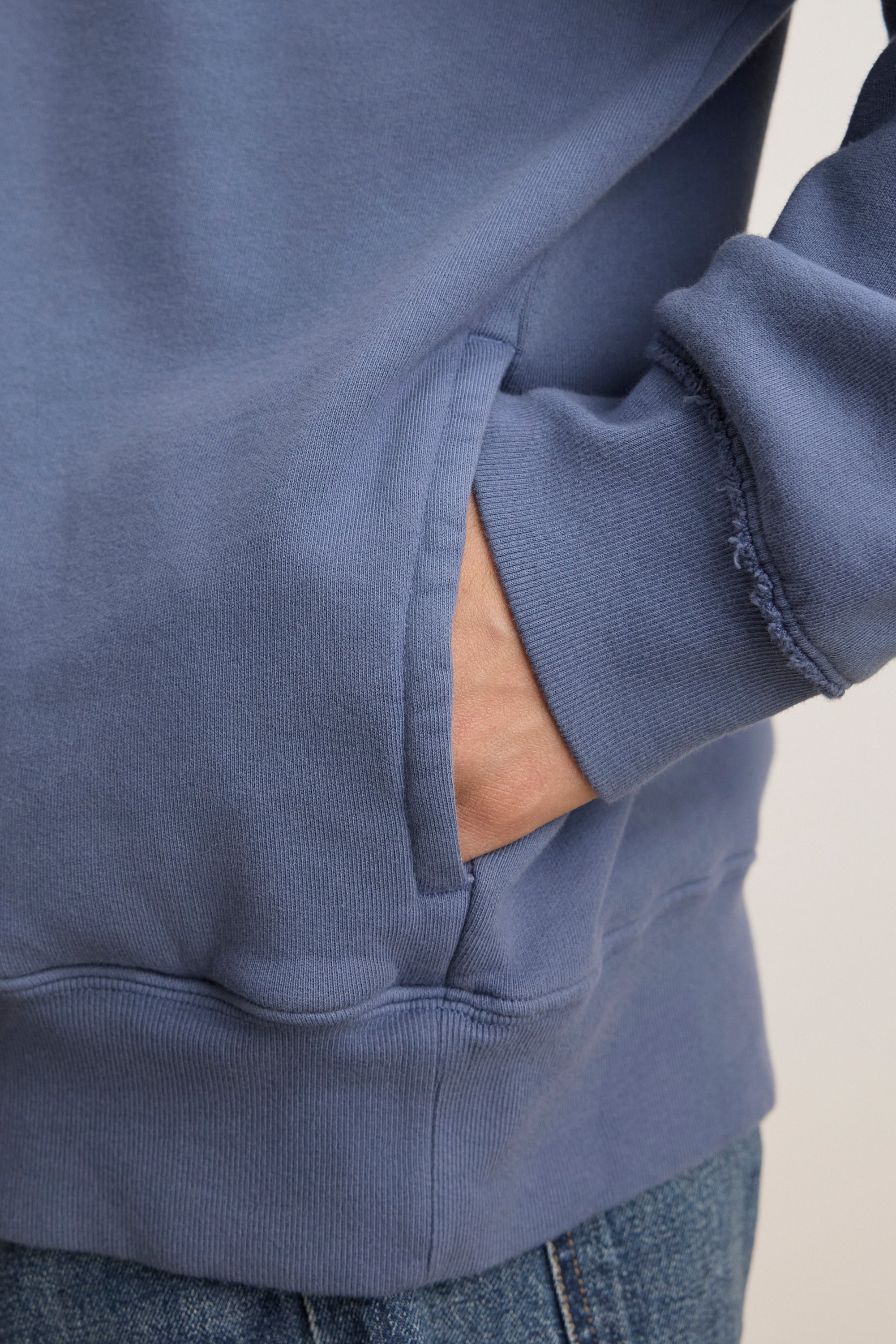   Someone wearing the Velvet by Graham & Spencer's FORREST SWEATSHIRT in a relaxed fit blue design, showcasing frayed fabric near the sleeve with a hand in the pocket. 