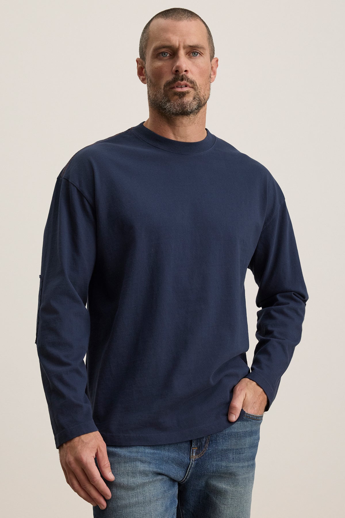 A short-haired, bearded individual stands against a neutral background wearing Velvet by Graham & Spencer's SLATER TEE, a dark blue long-sleeve pima cotton jersey, paired with jeans. It offers a relaxed fit suitable for any occasion.-38500088643777