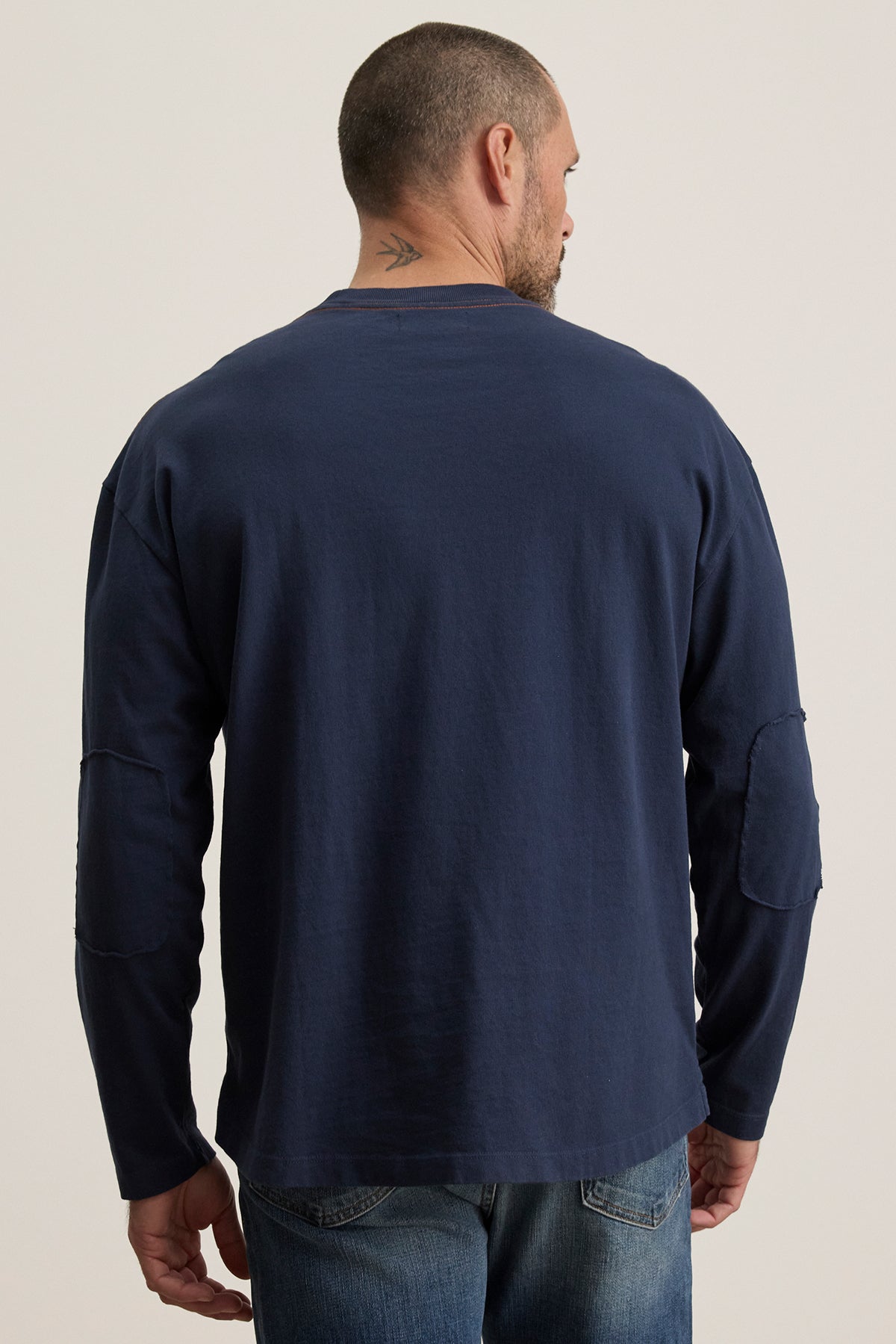   A man stands with his back turned, wearing Velvet by Graham & Spencer's SLATER TEE in navy, paired with blue jeans. The relaxed fit ensures comfort, and a tattoo on the back of his neck adds an element of intrigue. 