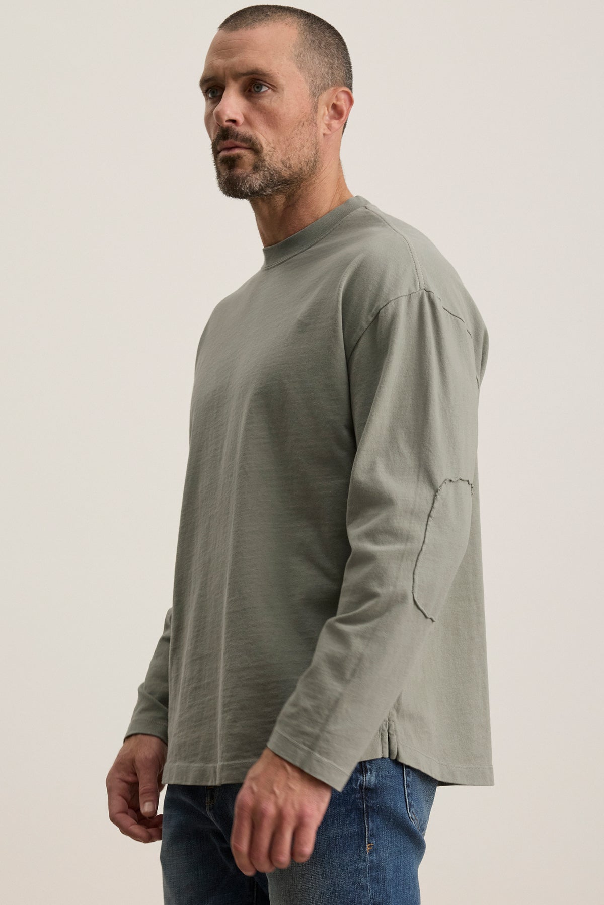   Wearing the SLATER TEE, a relaxed-fit green long-sleeve shirt made of structured pima cotton from Velvet by Graham & Spencer, a man stands against a neutral background, showcasing versatile style paired with jeans. 