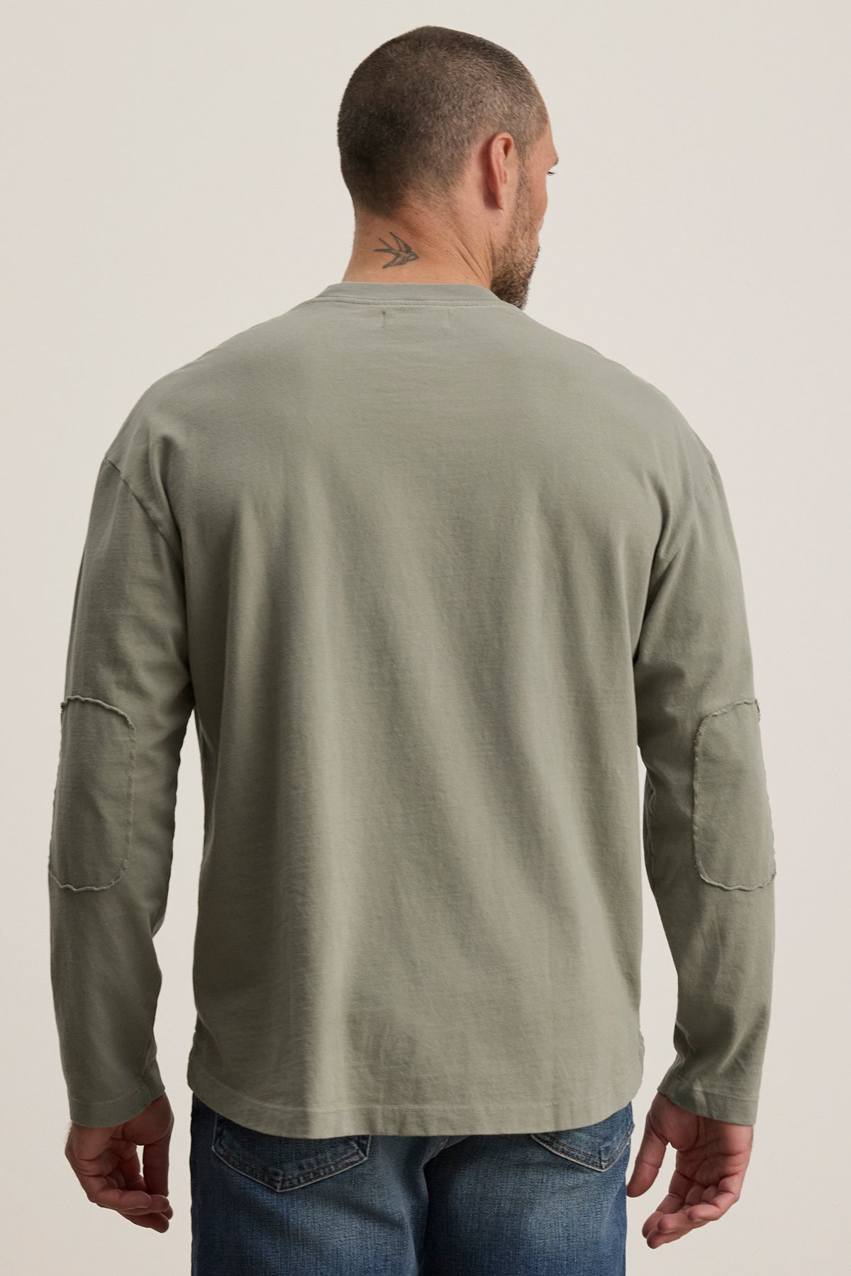   A man with a short haircut and neck tattoo wears the Velvet by Graham & Spencer SLATER TEE, a gray structured pima cotton long-sleeve with elbow patches. Paired with blue jeans, he stands facing away from the camera. 