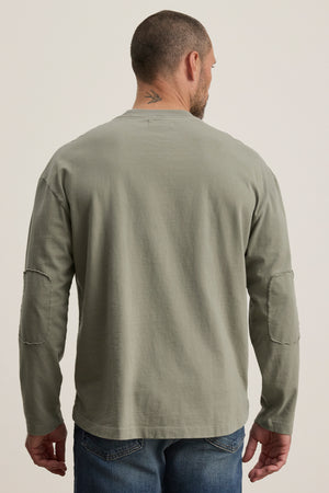 A man with a short haircut and neck tattoo wears the Velvet by Graham & Spencer SLATER TEE, a gray structured pima cotton long-sleeve with elbow patches. Paired with blue jeans, he stands facing away from the camera.