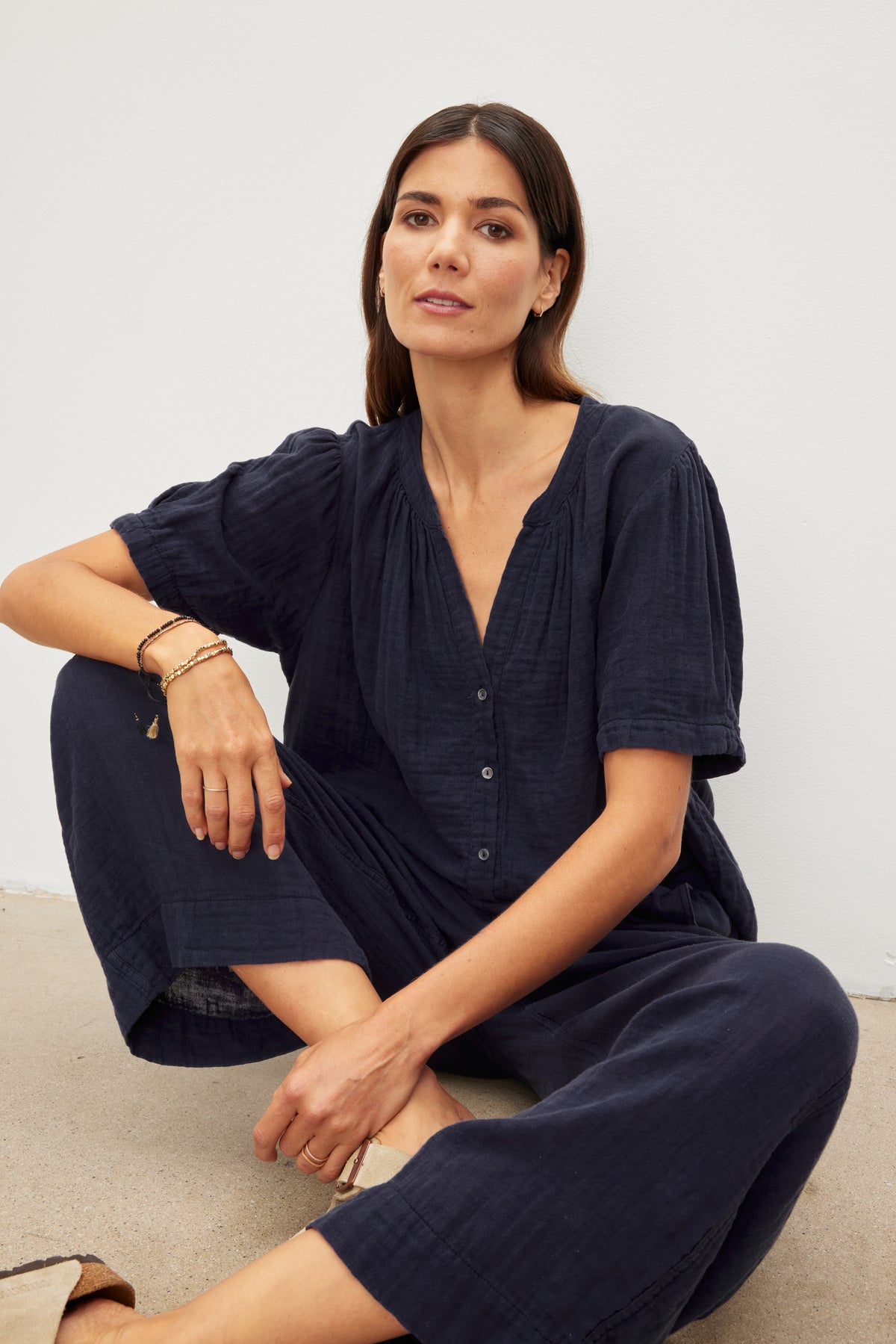   Woman in a casual navy blue outfit featuring FRANNY COTTON GAUZE PANTS by Velvet by Graham & Spencer with relaxed legs, sitting against a neutral background. 