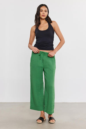 A woman stands confidently, wearing black tank top and Velvet by Graham & Spencer FRANNY COTTON GAUZE PANTS with relaxed legs and slash pockets, paired with black sandals. She's posing in a plain studio setting.