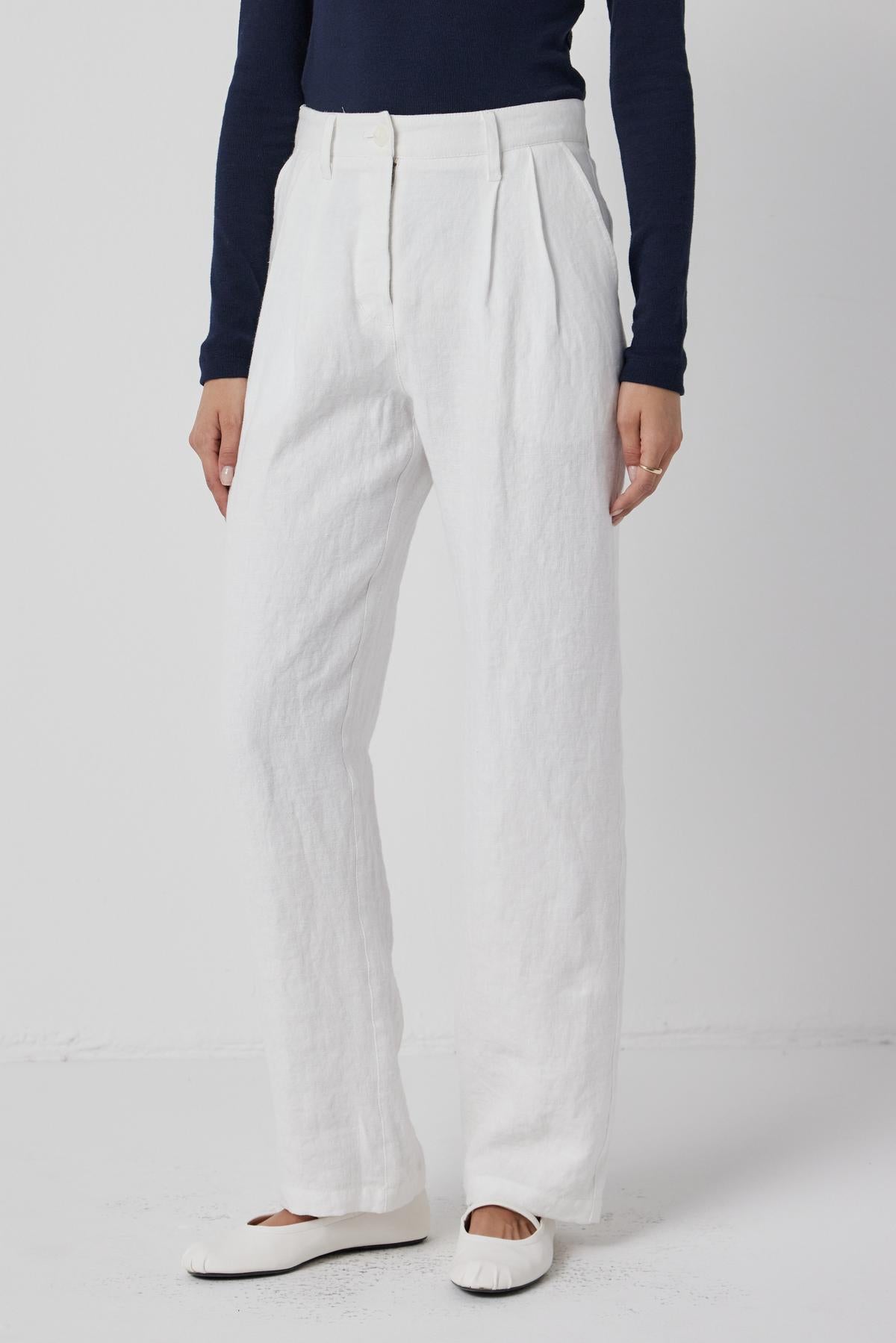   A person standing in Velvet by Jenny Graham's POMONA PANT - white straight-leg linen trousers with pockets, paired with a dark top and white flat shoes. 