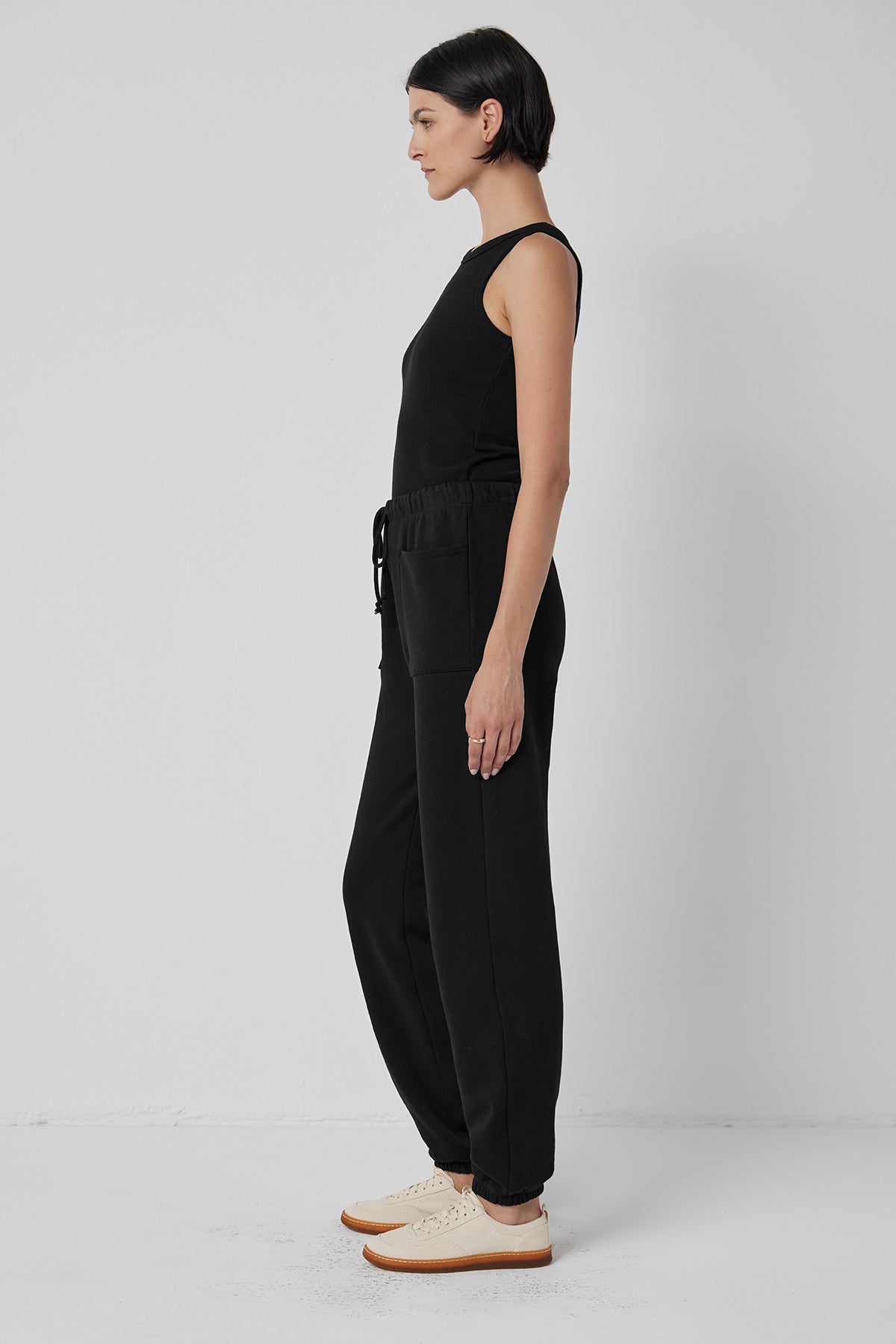   Person in a WESTLAKE SWEATPANT by Velvet by Jenny Graham, paired with a black sleeveless jumpsuit featuring an elastic drawstring waist, and white shoes, standing sideways against a plain white background. 