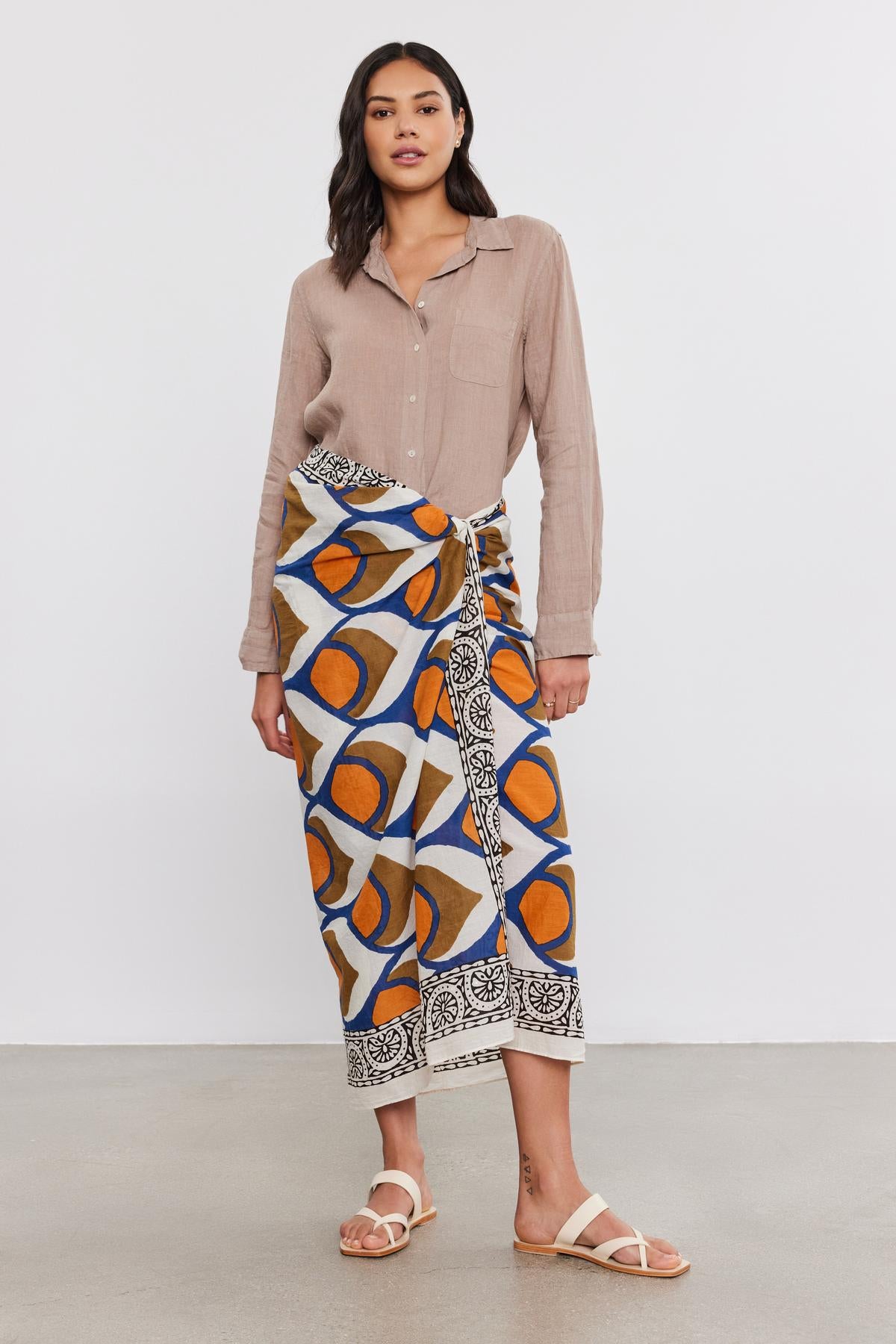   A woman wearing a beige shirt and a vibrant, patterned Velvet by Graham & Spencer sarong wrap with white sandals stands on a plain background. 