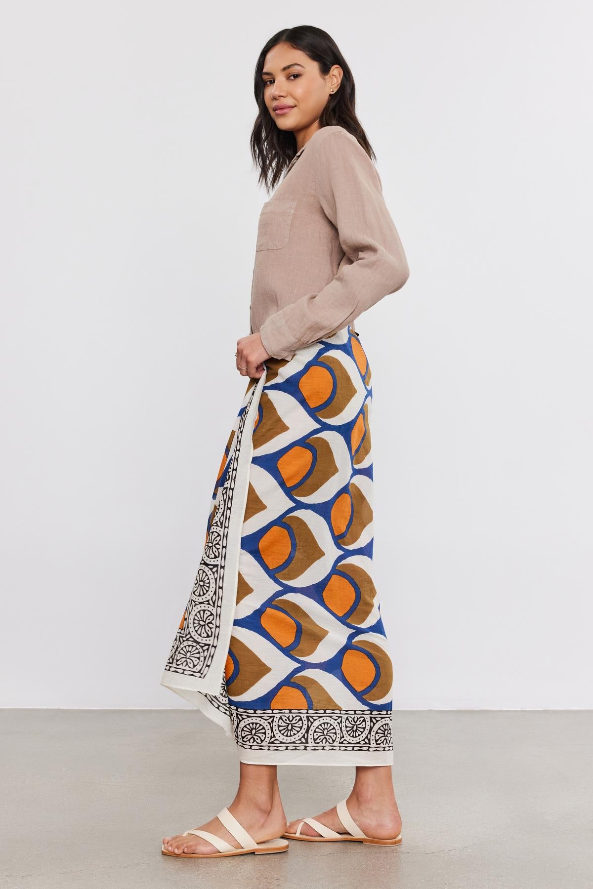   Woman standing sideways, wearing a beige blouse, colorful printed skirt, and white sandals by Velvet by Graham & Spencer, holding a SARONG WRAP bag, looking over her shoulder. 