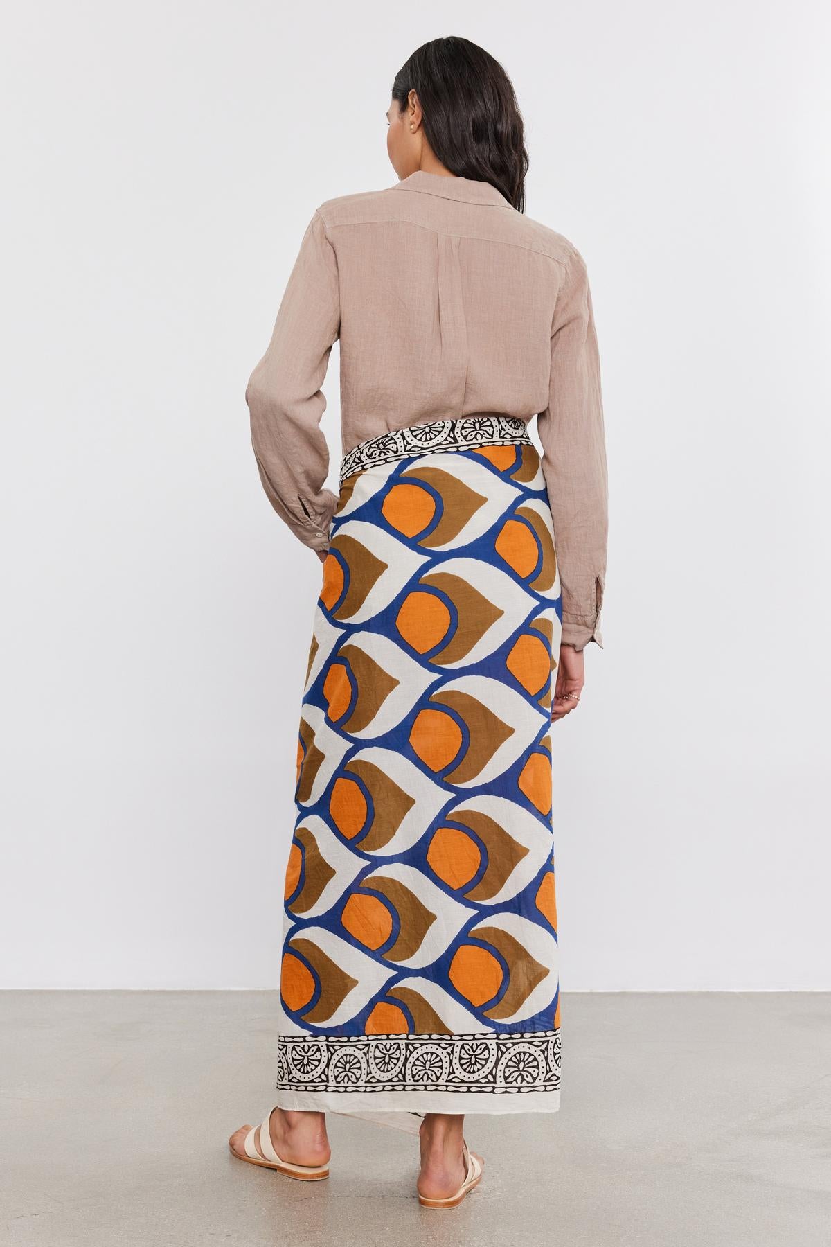   A woman standing with her back to the camera, wearing a beige blouse and a colorful geometric-patterned SARONG WRAP skirt from Velvet by Graham & Spencer, paired with tan sandals. 