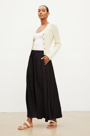 Woman posing in a white ribbed cardigan over a white tank top with a BAILEY LINEN MAXI SKIRT by Velvet by Graham & Spencer featuring an elastic drawstring waist, and beige sandals against a plain white background.