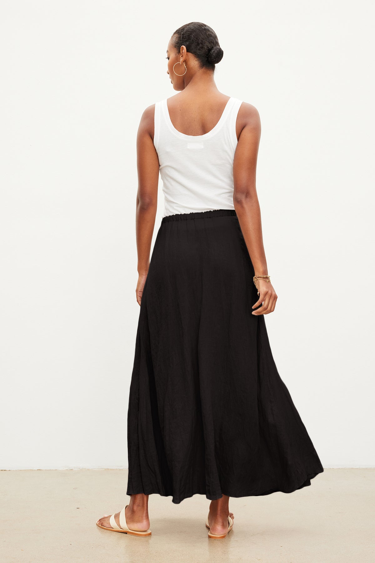   Person wearing a white sleeveless top, Velvet by Graham & Spencer BAILEY LINEN MAXI SKIRT with an elastic drawstring waist, and sandals, standing with their back facing the camera against a plain background. 