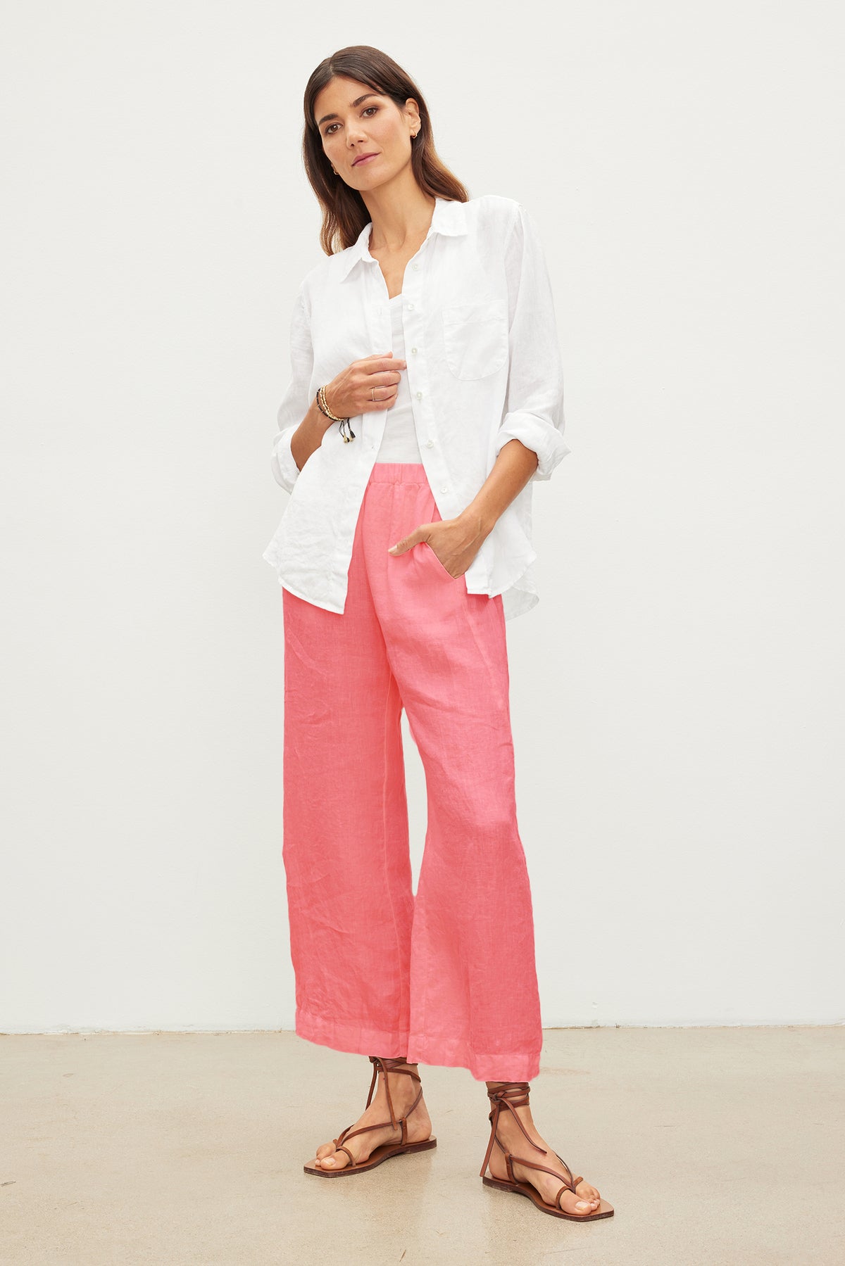   A woman stands against a plain white background, wearing a white button-up shirt and pink Velvet by Graham & Spencer LOLA LINEN PANT with an elastic waist. She has brown hair and is wearing sandals. 