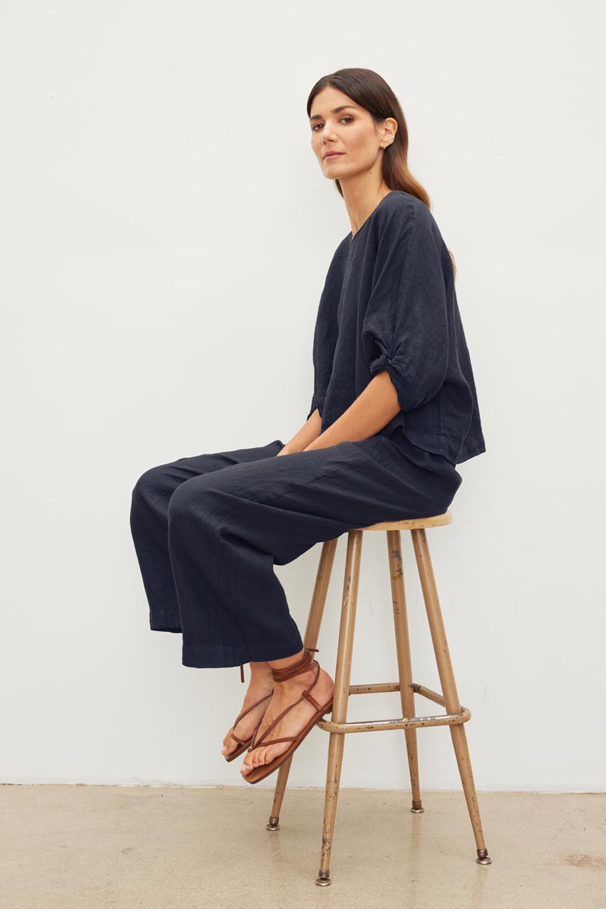   Woman with long dark hair sits on a wooden stool wearing a loose-fitting navy outfit with Velvet by Graham & Spencer's LOLA LINEN PANT and brown sandals, against a plain white background. 