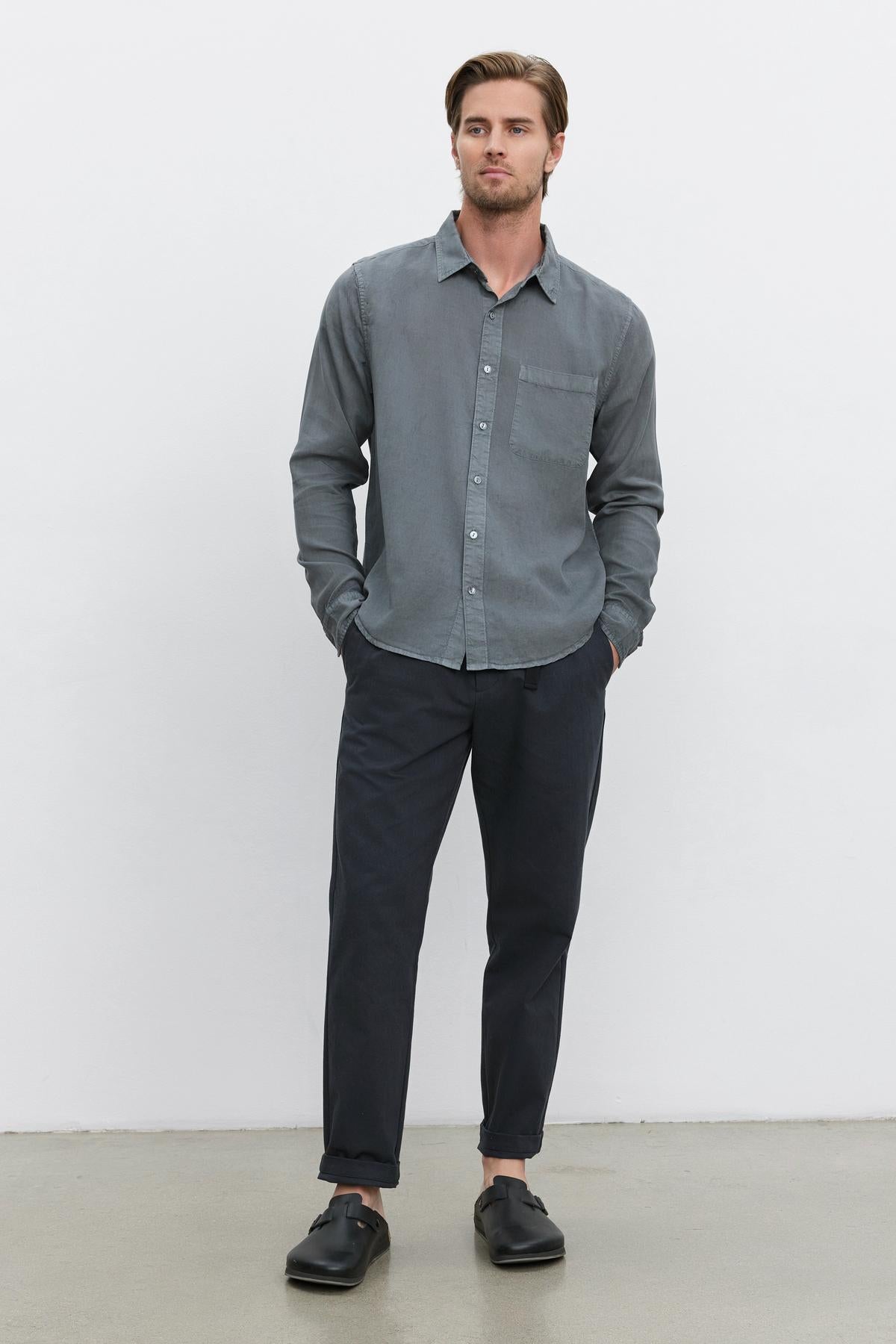 A man stands against a plain white background wearing the WOLF BUTTON-UP SHIRT by Velvet by Graham & Spencer, along with dark pants and black shoes, with his hands in his pockets.-37643215044801