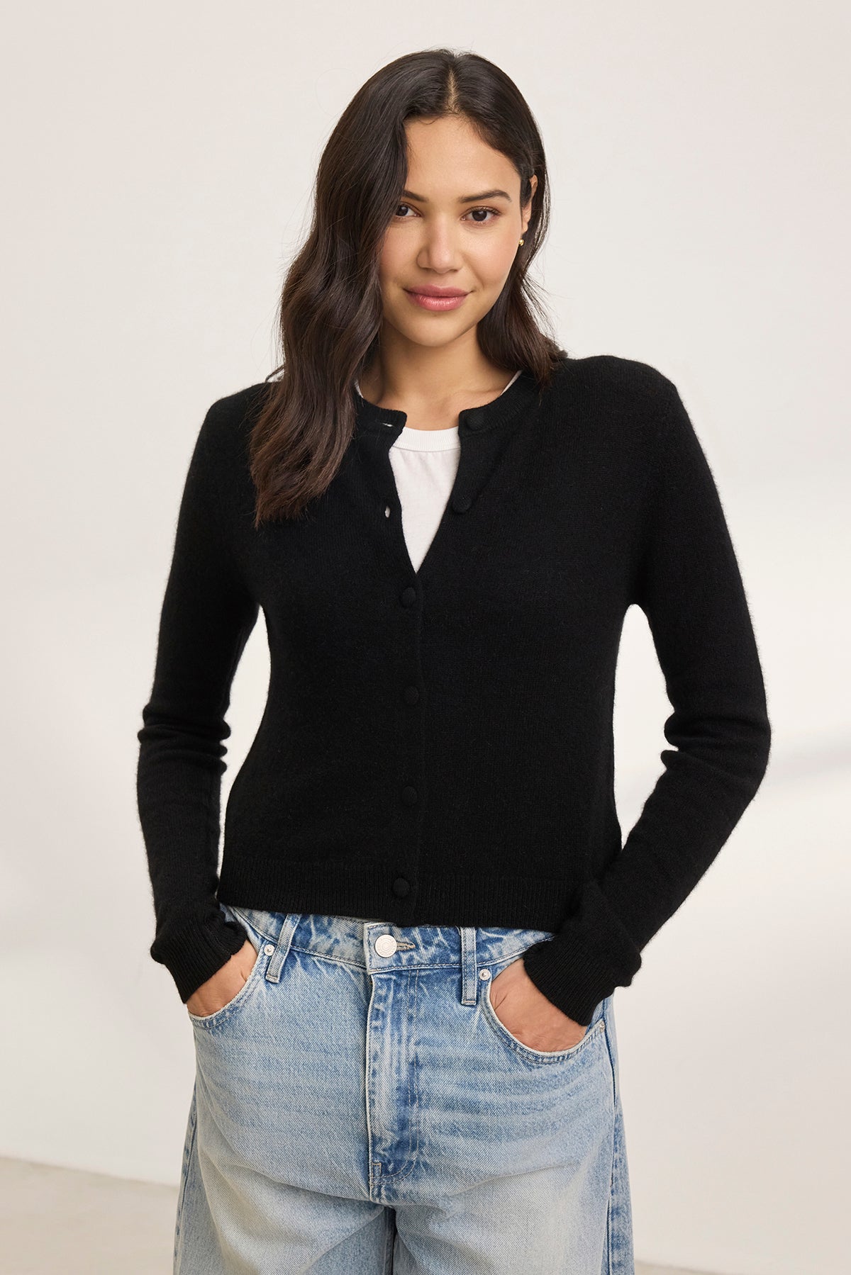   A woman stands against a plain background, wearing the CHRICHET CASHMERE CARDIGAN from Velvet by Graham & Spencer over a white shirt, paired with blue jeans. 