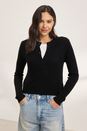 A woman stands against a plain background, wearing the CHRICHET CASHMERE CARDIGAN from Velvet by Graham & Spencer over a white shirt, paired with blue jeans.