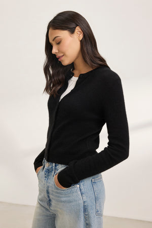 A woman wearing the CHRICHET CASHMERE CARDIGAN by Velvet by Graham & Spencer and blue jeans stands with hands in pockets against a plain background.