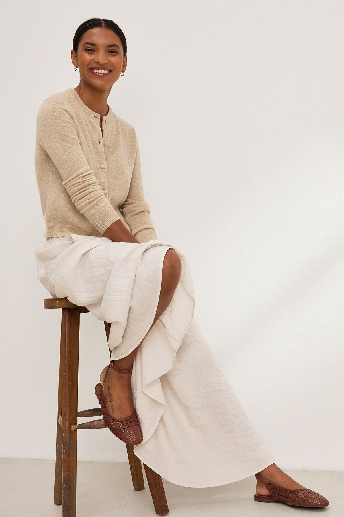   A person on a stool smiles against a plain background, wearing Velvet by Graham & Spencer's CHRICHET CASHMERE CARDIGAN in beige over a white skirt, paired with button-front brown woven shoes. 