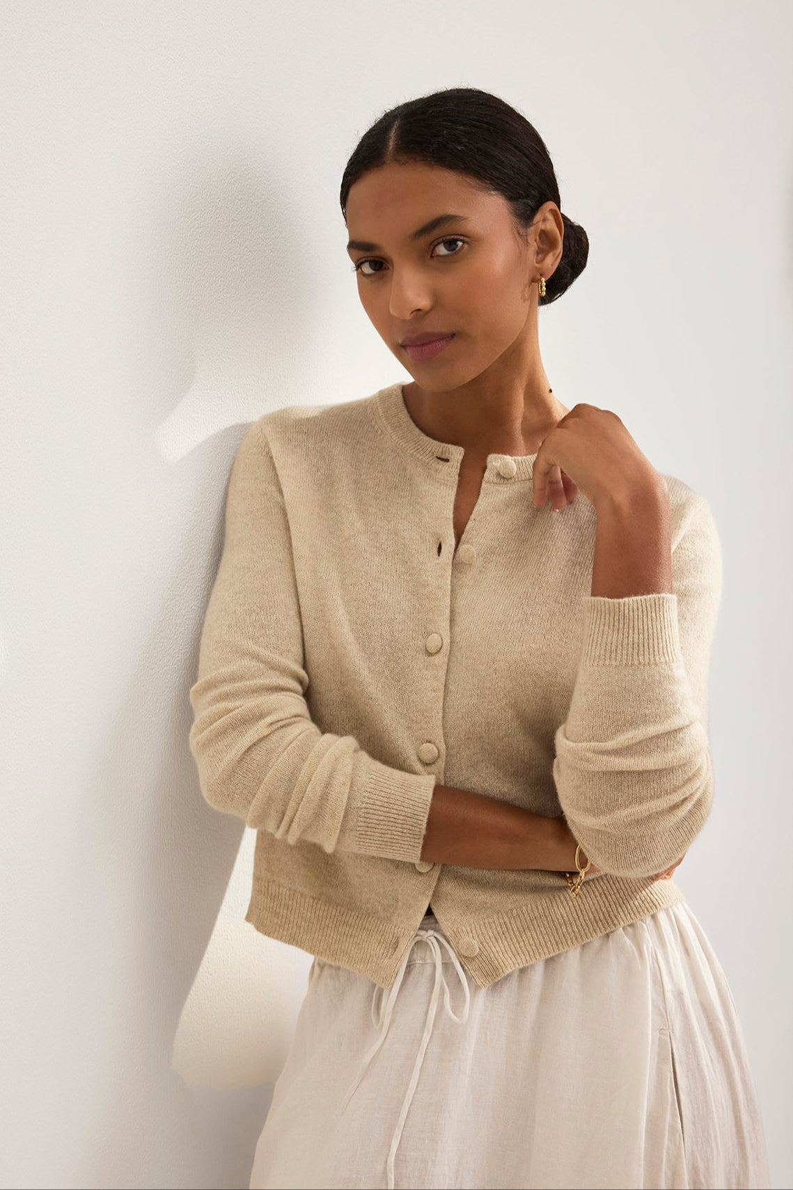   A person with dark hair in a low bun wears the CHRICHET CASHMERE CARDIGAN by Velvet by Graham & Spencer and a white skirt. They stand against a light-colored wall, gazing forward with one hand touching their neck. 