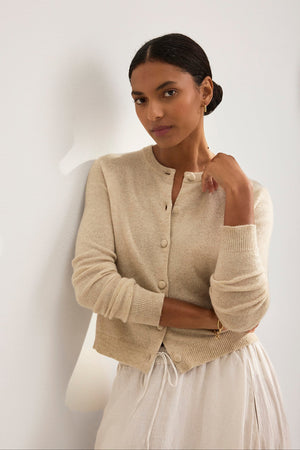 A person with dark hair in a low bun wears the CHRICHET CASHMERE CARDIGAN by Velvet by Graham & Spencer and a white skirt. They stand against a light-colored wall, gazing forward with one hand touching their neck.