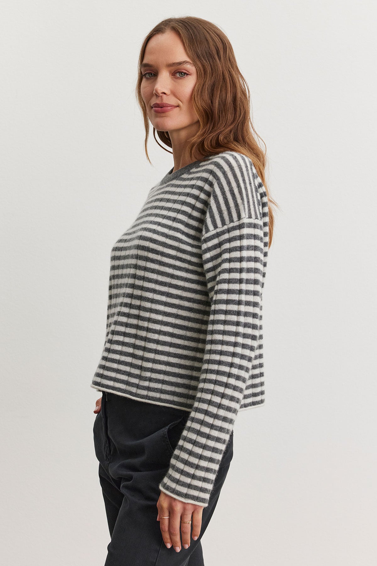   A woman wearing the MONICA CASHMERE SWEATER by Velvet by Graham & Spencer, featuring gray and white stripes, paired with dark pants, stands against a plain white background. 