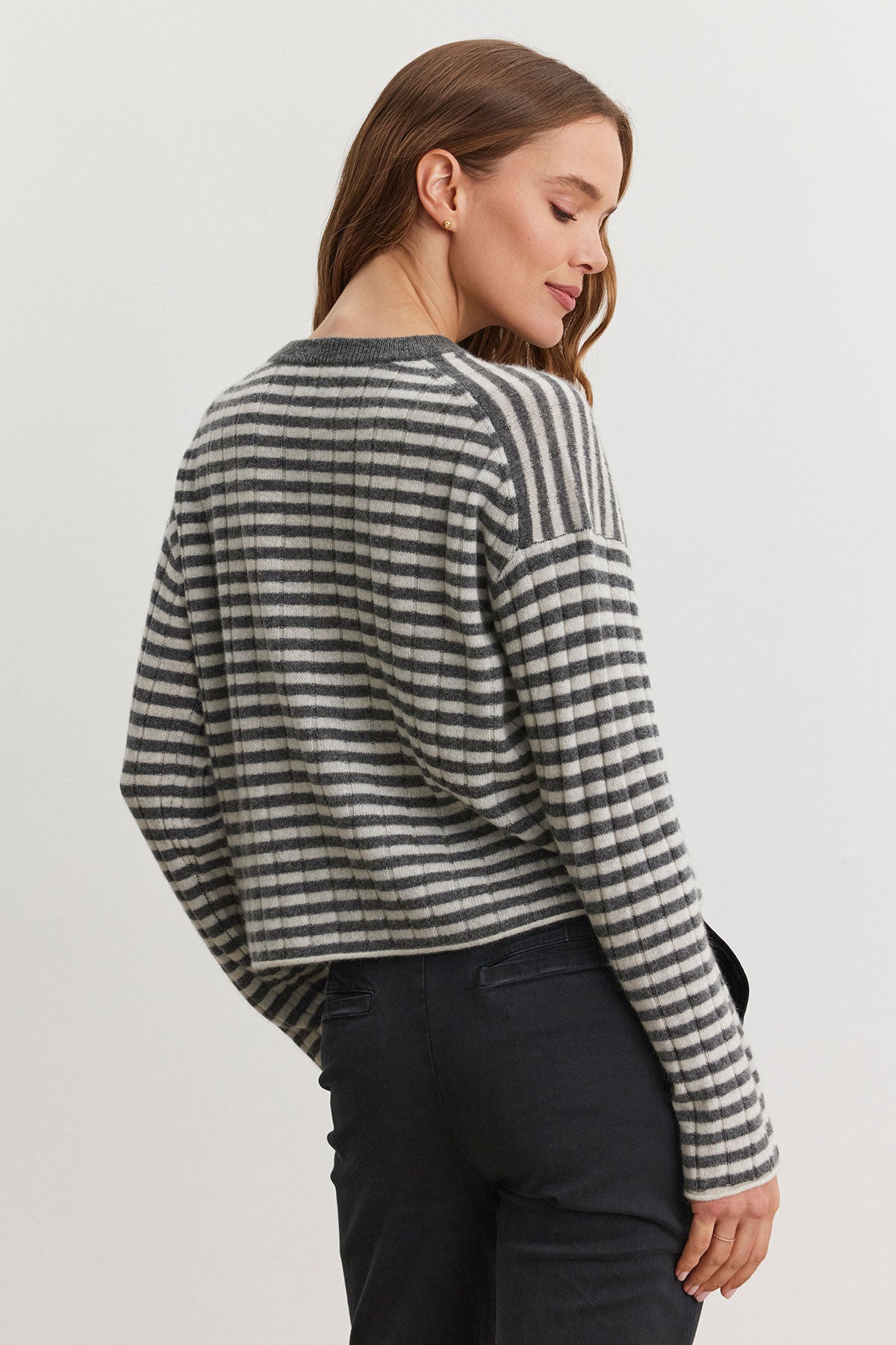 A person wearing the MONICA CASHMERE SWEATER by Velvet by Graham & Spencer, a wardrobe essential for luxurious comfort in black and white stripes, along with black pants stands against a plain background, facing slightly away.-38301405348033