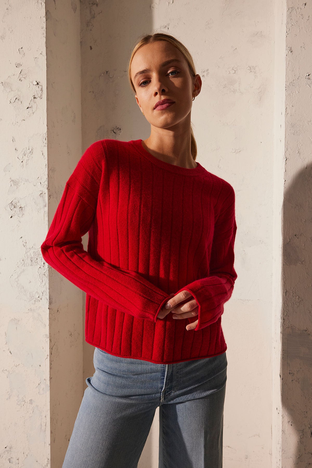   A person wearing the MONICA CASHMERE SWEATER by Velvet by Graham & Spencer, paired with blue jeans, stands against a textured white wall, showcasing this wardrobe essential. 