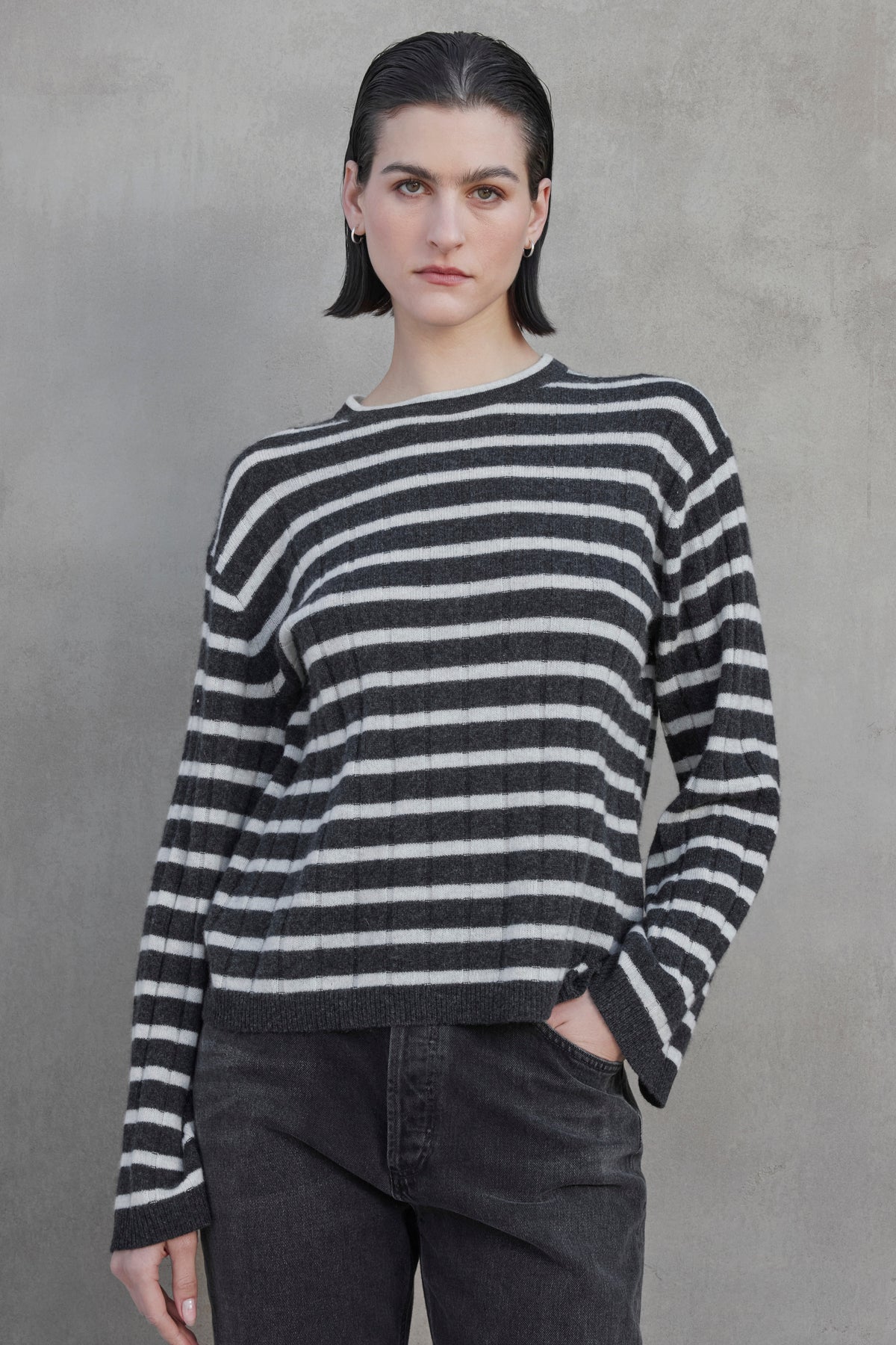   A person with short hair is standing against a gray wall, wearing a black and white striped NAPA CASHMERE SWEATER by Velvet by Jenny Graham and dark pants, with one hand in their pocket. 