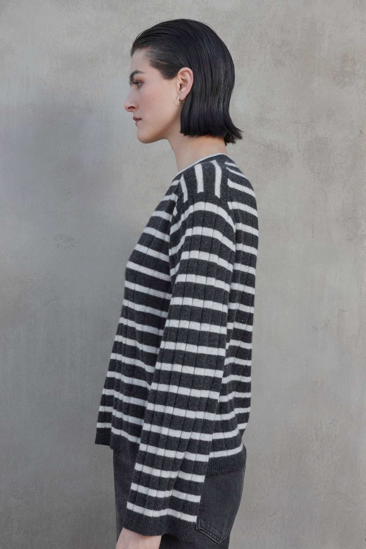   A side view of a person with short, dark hair wearing the NAPA CASHMERE SWEATER by Velvet by Jenny Graham, standing against a plain, gray textured wall. 